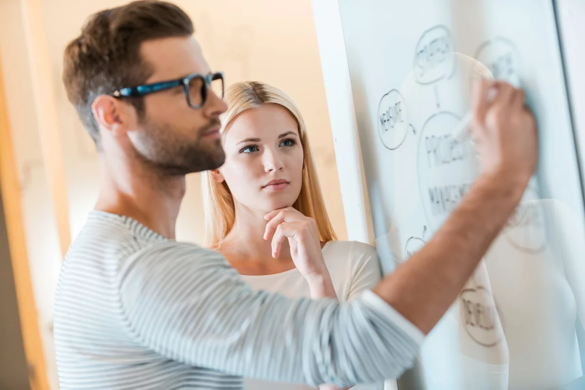 A young male developer is writing on a whiteboard as a blonde woman standing next to him watches.